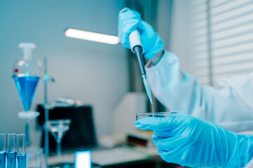 Woman in a lab coat is holding a pipette and pouring a blue liquid into a beaker