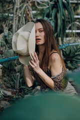 Obraz premium A stylish young woman in a cowboy hat poses in a lush greenhouse with exotic plants. She wears a leopard print shirt, beige shorts, and brown sandals, creating a casual safari-inspired look.