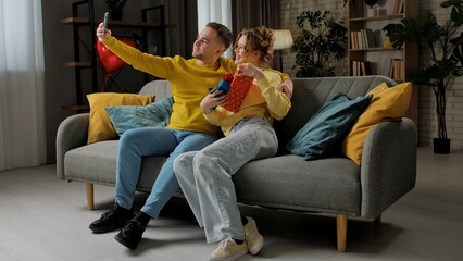 Obraz premium Happy couple celebrating valentines day at home, man and woman sitting on the sofa, hugging and taking selfie on smartphone with gifts, happy expression.