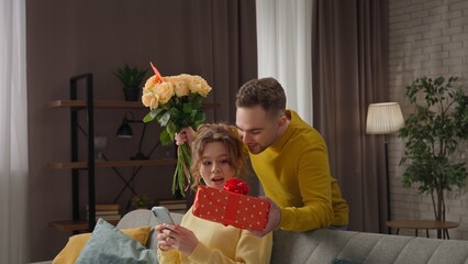 Happy couple celebrating valentines day together at home, woman sits on the sofa, man surprising girl with gift and flowers, amazed expression.