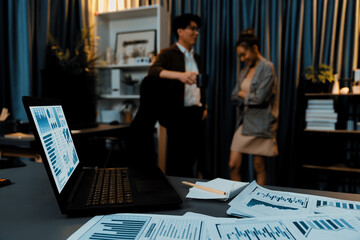 Setting on laptop showing business graph surrounded by paperwork marketing planning project on desk against on managers talking together background at over late night time at modern office. Infobahn.