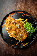 A beautifully plated crispy fried fish on a bed of golden French fries, garnished with fresh greens and drizzled with a tangy sauce, presented on a dark plate.
