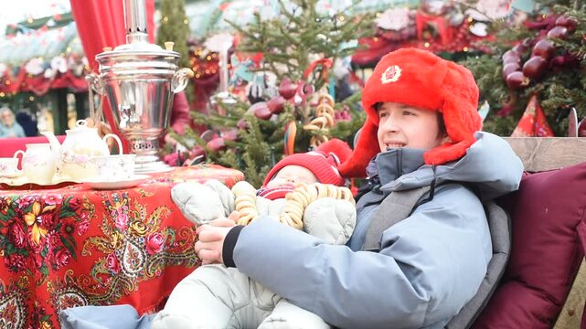  A boy in a red ushanka hat with the USSR coat of arms holds his sleeping baby brother, the baby brother has beads made of pretzels. Children sit at a table with a samovar on Maslenitsa. Pancake week.
