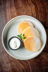 Three fluffy pancakes dusted with powdered sugar are served on a white plate, accompanied by a small black bowl of creamy yogurt and a fresh mint leaf.