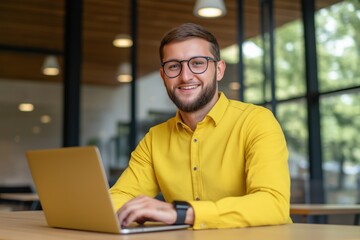 Fototapeta premium A man in a yellow shirt is sitting at a table with a laptop in front of him. He is smiling and he is enjoying himself