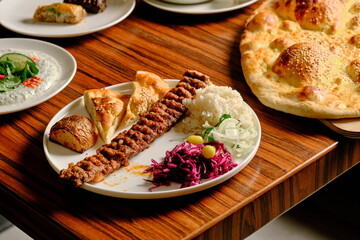Adana kebab with rice, red cabbage, potatoes, and bread on a plate, with Turkish balloon lavash bread beside it.