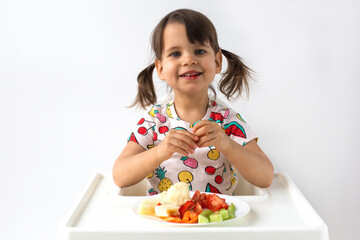 smiling little girl with pigtails sits in a high chair eating fresh fruits and vegetables. She wears a colorful t-shirt with fruit prints and looks happy. Concept of healthy eating and childhood