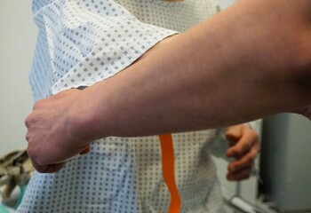 Arm von Ärztin beim Anlegen von Band am Arm von sitzender Patientin für Infusion im Krankenhaus 