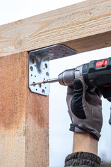A person uses a power drill to attach a metal bracket to wooden beams. Protective gloves are worn,...