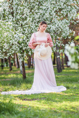 Fototapeta premium Full-length portrait of a young woman wearing a pink dress and hat posing against the backdrop of a spring park and white blooming apple trees.