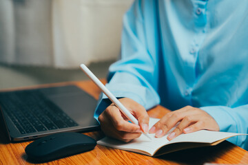 Person Writing in Notebook with Stylus Next to Laptop on Wooden Desk, Business concept.
