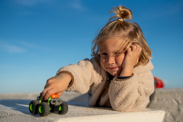 Thoughtful Child Lying on a White Concrete Surface, Playing with a Toy Monster Truck Under a Clear Blue Sky © sutulastock