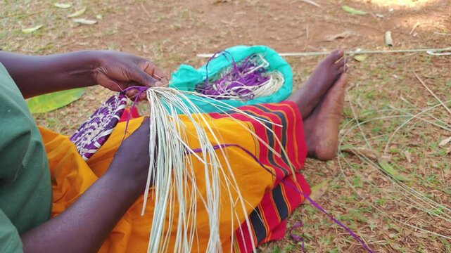 Person weaving colorful materials by hand while sitting on the ground. Uganda traditional craft