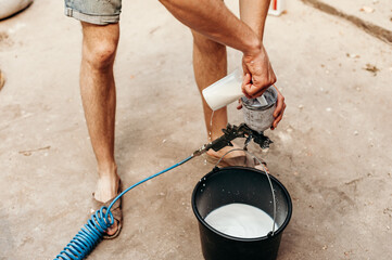 A man prepares a spray gun for work, pouring white paint into a container against the background of...