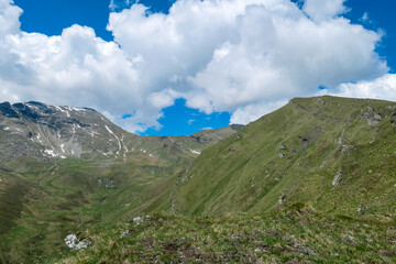Fototapeta premium Rocky cliffs meet lush, green valley near peak Reitereck, Austria. Lieser-Maltatal valley stretches out, framed by the distant, snow-capped Ankogel Group mountains, creating a scenic alpine landscape