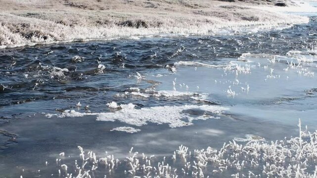 Fast-moving stream of narrow river runs along frozen banks on sunny day. Landscape of autumn nature with melting ice islands and frosted grass