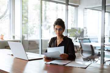 Focused latin hispanic young business woman working on laptop computer reading financial document report in office. Accountant entrepreneur manager businesswoman doing paperwork using pc. Copy space