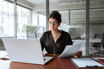 Focused latin hispanic young business woman working on laptop computer reading financial document report in office. Accountant entrepreneur manager businesswoman doing paperwork using pc. Copy space