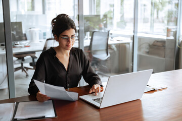 Focused latin hispanic young business woman working on laptop computer reading financial document report in office. Accountant entrepreneur manager businesswoman doing paperwork using pc. Copy space