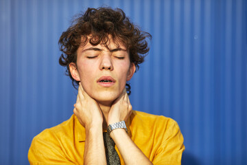 Young male student with curly hair expressing deep thought against a blue background while dressed...