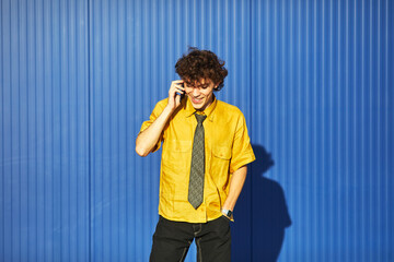 Young student in yellow shirt talking on the phone against a blue wall during the day