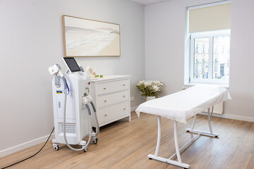 A clean room with a white treatment table near a window, a medical device with a touchscreen, a white dresser with gold knobs, and wooden flooring.