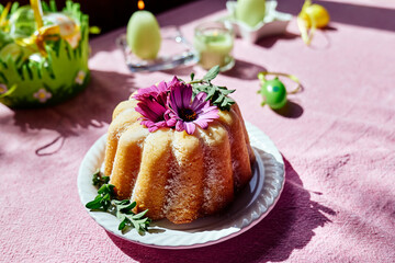 Easter cake decorated with icing sugar and daisy flowers on the table with easter eggs and tulips on  pink tablecloth. Traditional easter cake or sweet bread with topping. Easter treat.