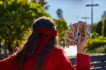 A woman in a red coat with long hair tied with a velvet bow holds five tarot cards in her hand...