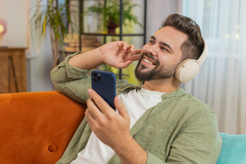Fototapeta premium Happy overjoyed young man in wireless headphones with smartphone relaxing lying on couch sofa at home apartment choosing, listening favorite energetic disco dancing music. Weekend leisure activities.