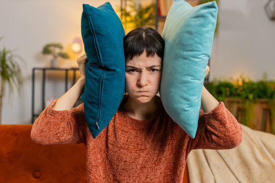Irritated Caucasian woman girl covers ears with cushions, annoyed by noisy neighbors suffers from headache sitting on couch. Thin walls at home, flat without sound insulation, repair work concept.
