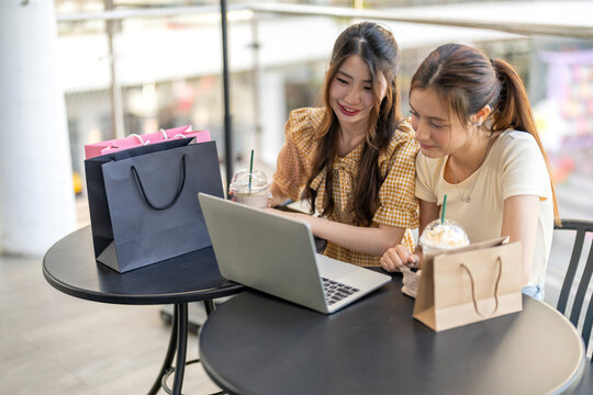 Two diverse women smiling and talking while use laptop for online shopping at cafe, searching for deals and discounts with shopping bags on table, concept of e-commerce, retail, digital lifestyle