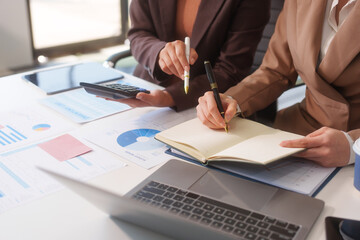 Close-up of two businesswomen working at desk discussing business strategies, analyzing financial charts, planning marketing plans and using laptop to drive business growth in professional office