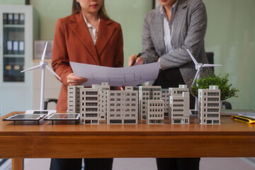 Two businesswomen working at desks and discussing sustainable urban development with emphasis on eco-friendly buildings,condos, wind turbines,solar cells achieving net zero carbon emissions better