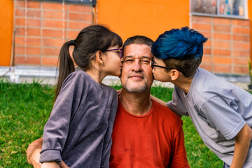 Obraz premium Photo of Colombian Latino grandfather looking into camera smiling with his two mestizo grandchildren kissing him on each cheek.