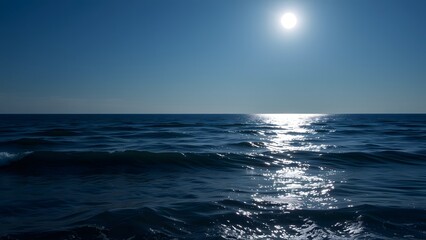 Obraz premium Serene ocean waves glistening under the radiant sun on a clear day, with the vibrant blue sky meeting the horizon for a peaceful and tranquil coastal scene.