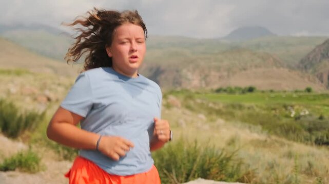 Exhausted overweight girl runs along a winding road through hills, struggling to lose weight under the scorching summer sun. Behind her, picturesque mountains rise beneath looming storm clouds.