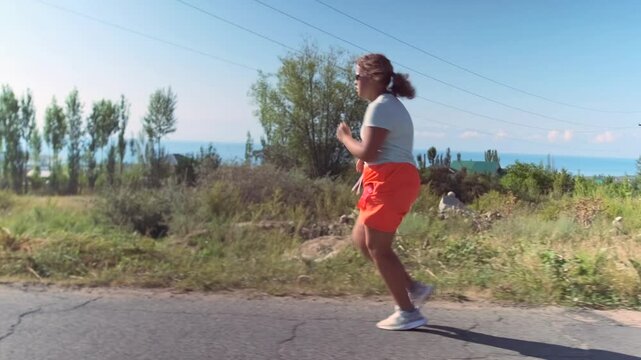 A young girl in a sportswear runs along the road, pushing through fatigue. Green shrubs and trees surround her, with a vast landscape under a clear blue sky.