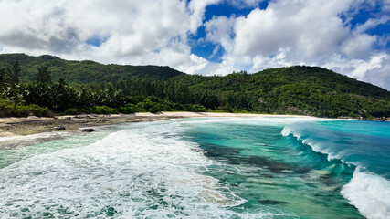 A large turquoise wave breaking near a tropical beach with soft sand, palm trees, and green hills. Seychelles, Mahe. Police Bay.