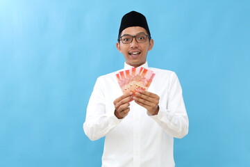 Wow excited asian muslim man standing while holding Indonesian banknotes. Isolated on blue