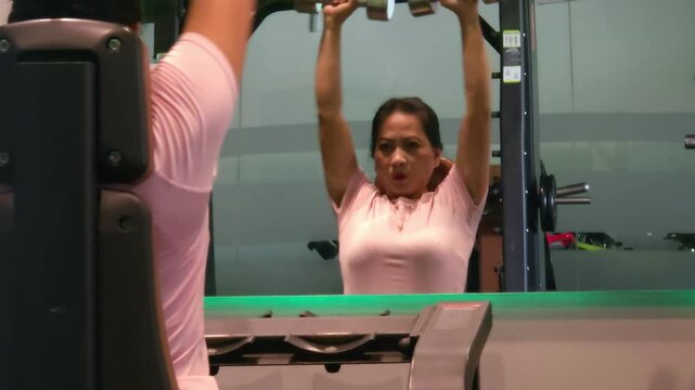Back view of 40 year old woman doing weightlifting in gym using dumbbells with blurry reflection in mirror.