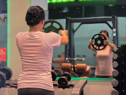 Back view of 40 year old woman doing weightlifting in gym using weight plates to train shoulder muscles with blurred reflection in mirror.