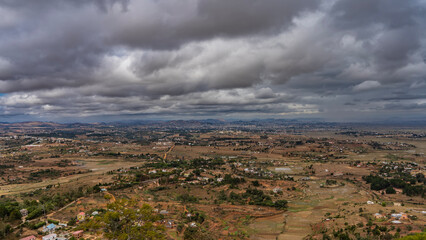 Rural landscape. View from the height. The valley has rural houses, green vegetation, and dirt roads. Agricultural fields are cultivated on red soil. Mountains on the horizon. Cloudy sky. Madagascar.