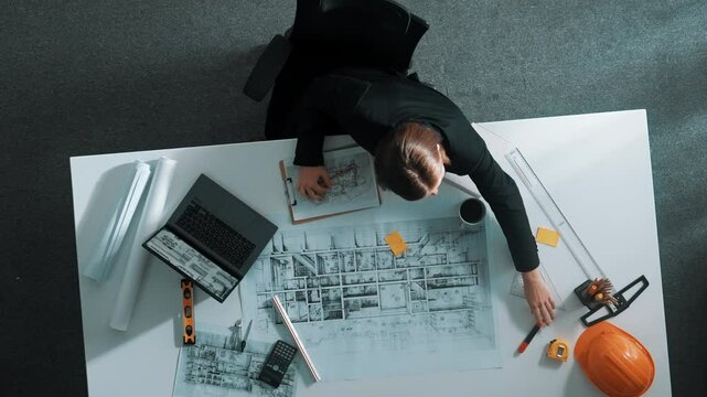 Aerial view of architect drawing while working at laptop. Top down view of caucasian engineer designing blueprint and drafting house model at table with equipment and safety helmet. Alimentation.