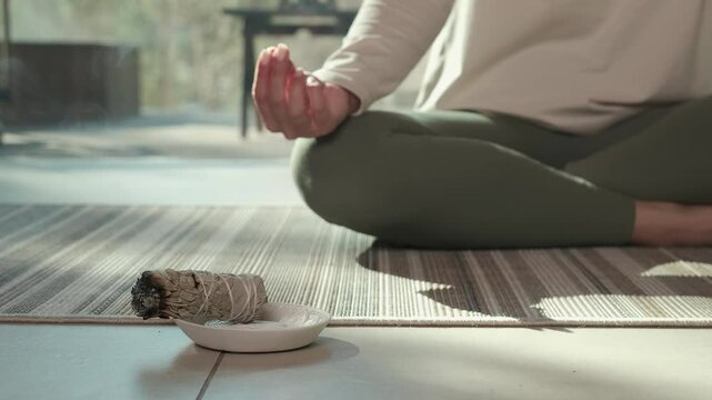 Sunlight illuminates a woman practicing yoga and meditation with her hands in a mudra position, while a smoldering white sage bundle rests on a small plate in front of her