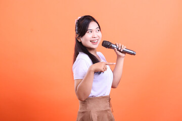 A cheerful young woman holds a microphone, smiling and pointing, ready to perform with passion. isolated on orange background