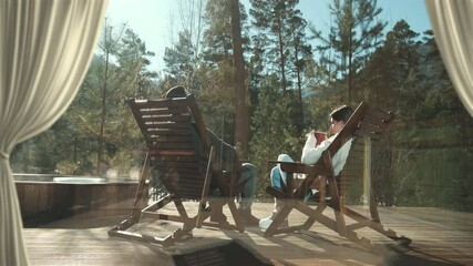 Sunlight streams through curtains onto a wooden deck where a couple relaxes in rocking chairs, enjoying the serene view of a steaming hot tub and a tranquil forest
