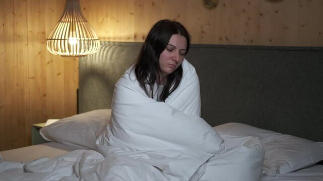 Woman sitting on bed wrapped in blanket feels cold and sick, suffering from fever or seasonal flu, experiencing discomfort and chills in bedroom at night