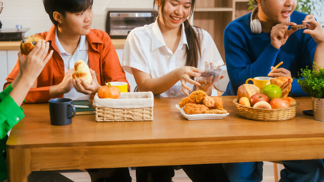 Five young Asian men and women gather around a kitchen desk, enjoying a party with drinks, fried chicken, and bread. They share laughter, good food, and happiness in a lively atmosphere.