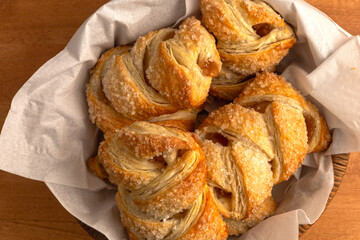 Close-up overhead view of golden-brown apple strudels with a crispy, flaky texture and a sugar-coated surface, placed in a small basket lined with white paper tissue on a wooden tabletop.