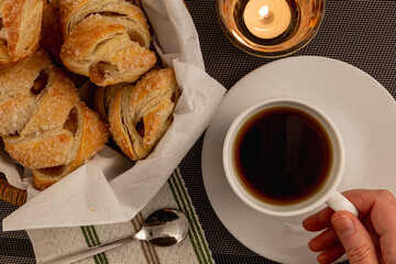 Overhead view of sugar-coated apple strudels,pastries, freshly baked and placed in a basket, with a cup of tea held by hand and a spoon on the napkin and lit candle nearby.